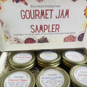 May include: A sampler box of six 4-ounce jars of gourmet specialty jams. The jars are labeled with the names of the jams: Caramel Apple, Monkey Jam, Rhubarb Crisp, Carrot Cake, Blueberry Muffin, and Pecan. The box is brown with white text and images of spices.