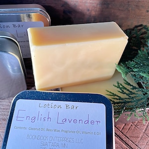 May include: A light yellow lotion bar with the text "English Lavender" on a label. The bar is rectangular and sits next to a metal tin and green sprigs. The label lists the ingredients.