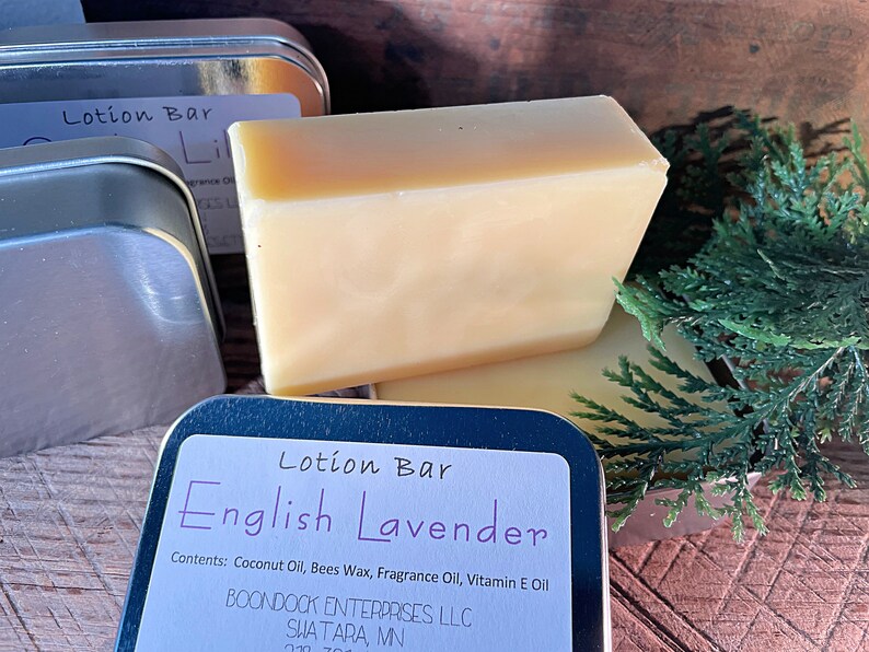 May include: A light yellow lotion bar with the text "English Lavender" on a label. The bar is rectangular and sits next to a metal tin and green sprigs. The label lists the ingredients.
