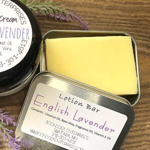 May include: A close-up of a lotion bar in a silver tin, labeled "English Lavender." The bar is a pale yellow color. A jar of lavender cream is visible, along with sprigs of lavender. The label lists ingredients.