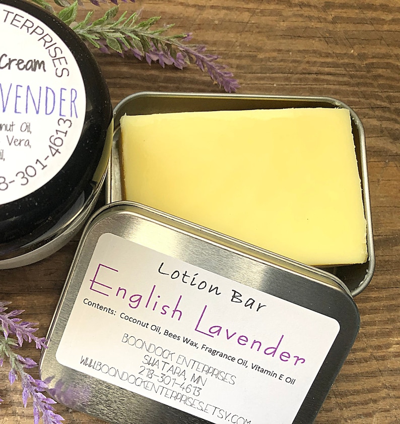 May include: A close-up of a lotion bar in a silver tin, labeled "English Lavender." The bar is a pale yellow color. A jar of lavender cream is visible, along with sprigs of lavender. The label lists ingredients.