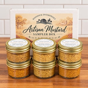 May include: A white cardboard box labeled Boondock Enterprises Artisan Mustard Sampler Box sits behind six small glass jars with gold lids. The jars contain textured mustard in Honey Pepper, Horseradish, and Sweet Thyme flavors with white labels.
