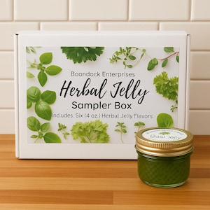 May include: A white box with the text "Boondock Enterprises Herbal Jelly Sampler Box" and images of herbs. Includes six 4 oz. jars of herbal jelly. A small jar of green basil jelly is in front of the box.