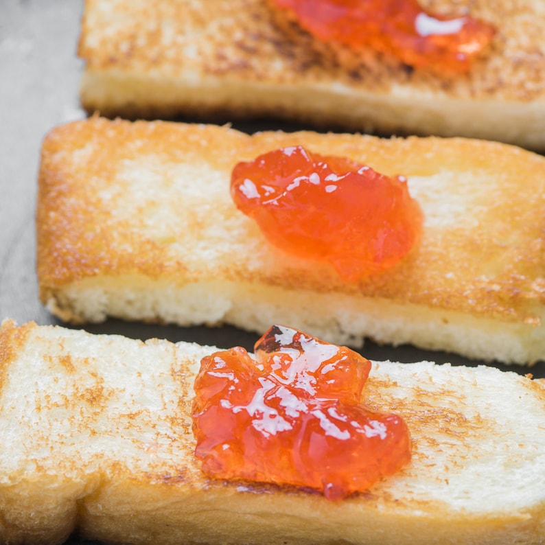 Puede incluir: Tres rebanadas rectangulares de pan tostado dorado dispuestas en fila. Cada rebanada de pan tiene una porci&oacute;n de mermelada de frutas de color naranja brillante, con una superficie brillante, texturizada y de aspecto ligeramente transl&uacute;cido.