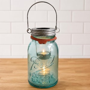 May include: A light blue glass Ball Perfect Mason jar repurposed as a lantern, featuring a silver metal lid with a matching wire handle, a rusted orange band around the neck, and a small lit tea light candle suspended inside the glass container.