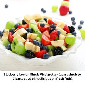 Op de afbeelding: Een witte kom gevuld met een kleurrijke fruitsalade. De salade bevat aardbeien, blauwe bessen, kiwi, druiven en bananen. De kom staat op een wit oppervlak.