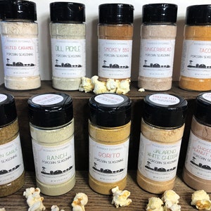 May include: A collection of 10 small glass jars with black lids filled with different colored powders. The jars are labeled with various popcorn seasoning flavors, including Salted Caramel, Dill Pickle, Smokey BBQ, Gingerbread, Taco, Parmesan Garlic, Ranch, Dorito, Jalapeno White Cheddar, and Chocolate Peanut Butter.