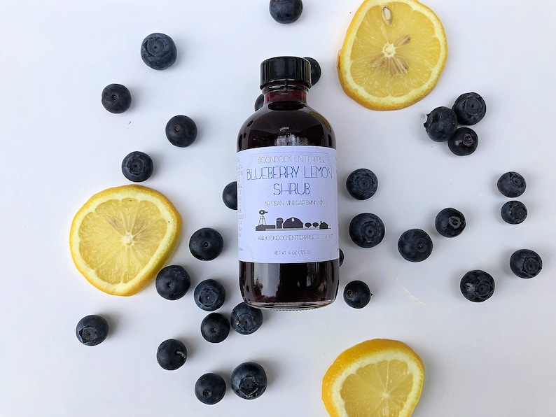 Op de afbeelding: Een glazen fles met bosbessen-citroensiroop, een ambachtelijke drankmixer op basis van azijn, omringd door bosbessen en citroenschijfjes op een witte achtergrond. Het etiket luidt "Boondock Entertainments Blueberry Lemon Shrub Artisan Vinegar Drink Mixer"