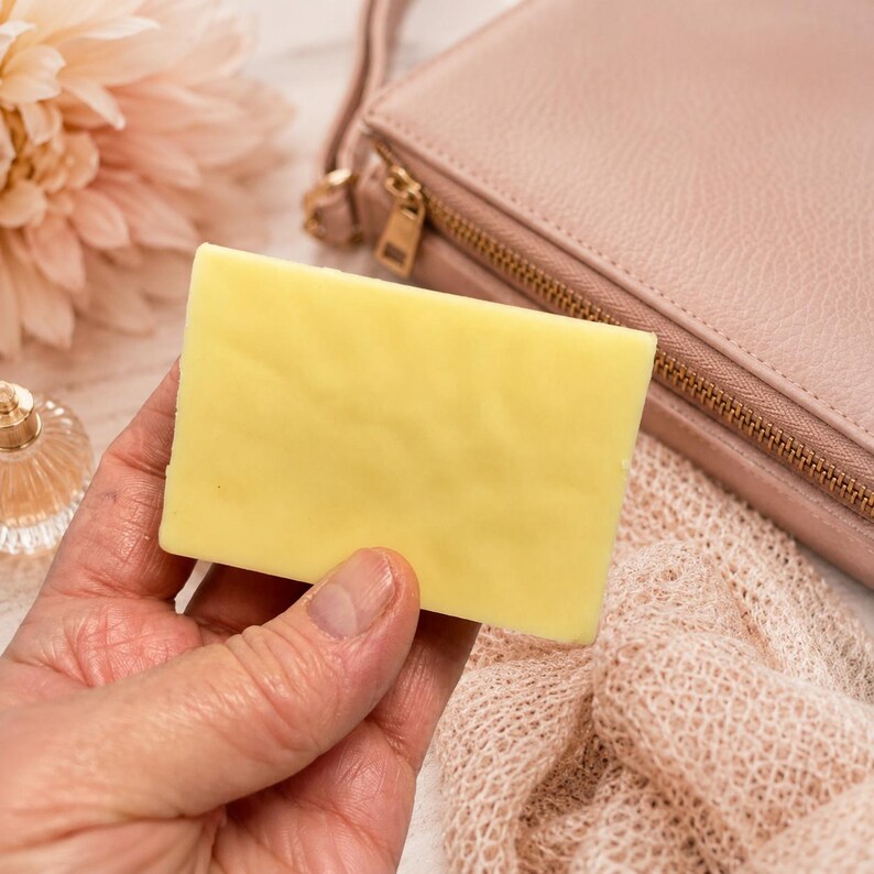 May include: A rectangular, pale yellow bar of soap is held in a hand. A pink purse with a gold zipper, a pink flower, and a small perfume bottle are in the background. The soap appears to be unscented.