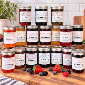 May include: A collection of eighteen glass jars filled with various fruit jams and jellies, each featuring a white label with black text. The jars are arranged in rows on a wooden surface, accompanied by fresh strawberries and blueberries.