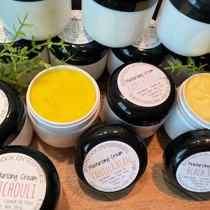 May include: A collection of small, white jars with black lids, each containing a different moisturizing cream. The jars are labeled with the name of the cream, "Boondock Enterprises", and the ingredients. The creams include "English Rose", "Vanilla Bean", "Baby Powder", "Patchouli", and "Black Spice".