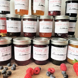 May include: A variety of homemade jams and marmalades in glass jars with labels. The jars are arranged in rows on a wooden surface. Some of the flavors include: Raspberry Plum, Jalapeno, Chokecherry, Strawberry, Cherry Jam, Orange Marmalade, and Rhubarb Jam.