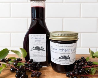 May include: A clear glass bottle of dark purple Chokecherry Syrup and a glass jar of dark purple Chokecherry Jelly, both with white labels featuring a house illustration, alongside fresh dark berries on stems on a wooden surface.