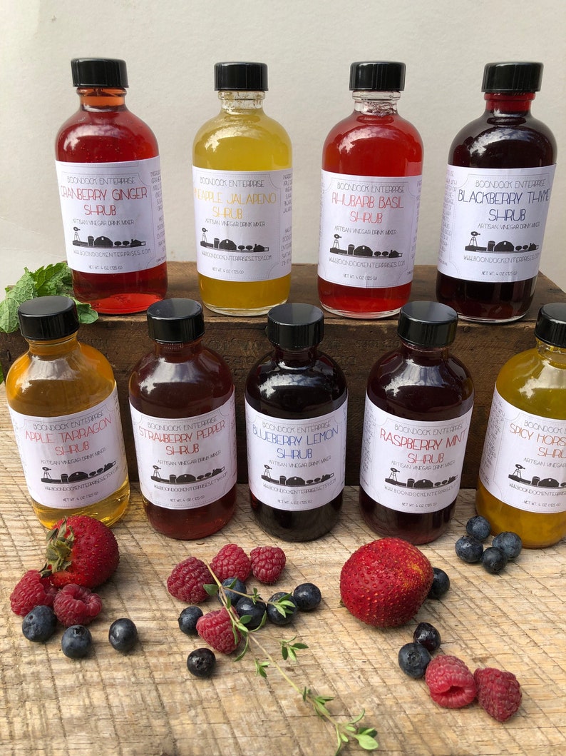 Op de afbeelding: Tien glazen flessen met verschillende kleuren en smaken van zelfgemaakte fruitsap, een drank op basis van azijn. De flessen zijn in twee rijen op een houten oppervlak gerangschikt, met verse bessen en kruiden die eromheen verspreid zijn. De etiketten op de flessen tonen de merknaam "Boondock Enterprises" en de smaak van elke sap.