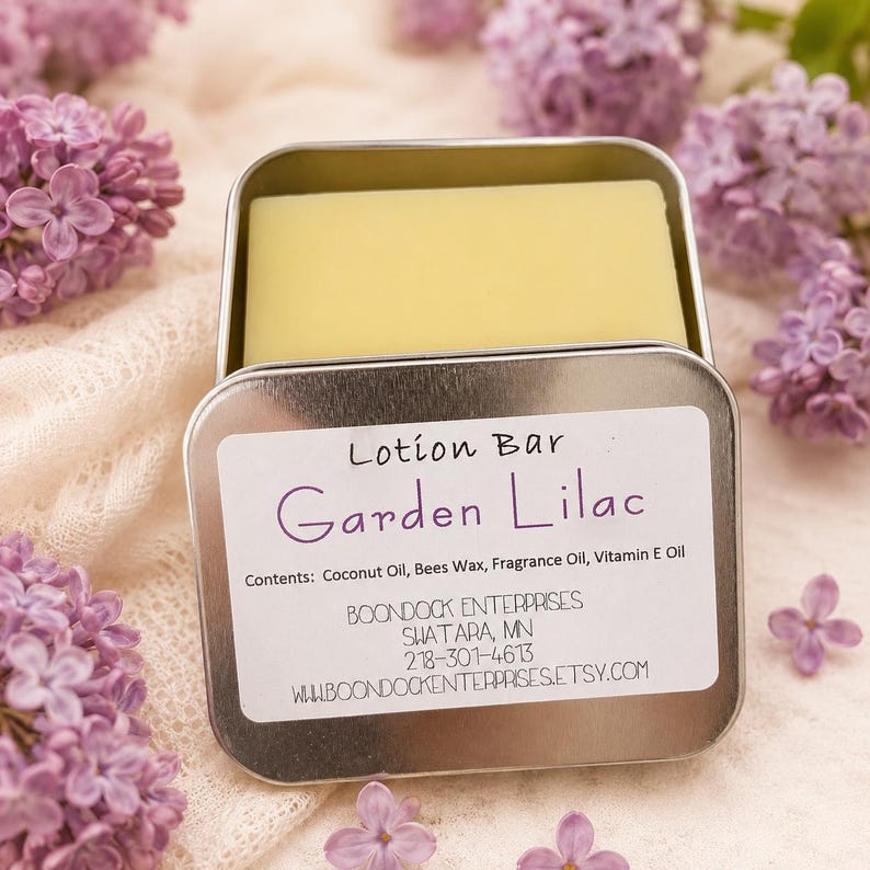 May include: A rectangular, pale yellow lotion bar sits inside an open, silver tin. The label reads "Lotion Bar Garden Lilac." The background features soft, white fabric and sprigs of purple lilac flowers.