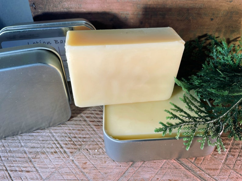 May include: Two rectangular lotion bars, one atop the other, with a pale yellow hue, are displayed. They rest in a small, silver tin. A sprig of green foliage adds a natural touch. The background is a wooden surface.