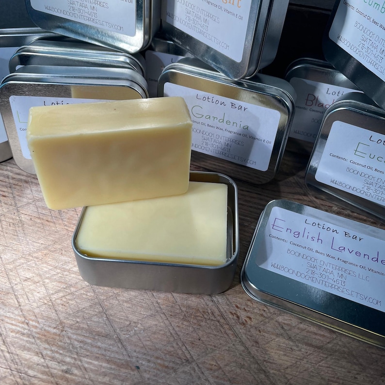 May include: A light yellow lotion bar sits in a small, silver tin, with another bar on top. Several other tins with labels are in the background. The labels read "Gardenia" and "English Lavender".