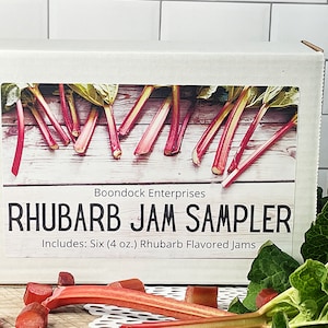 May include: A white box featuring a label with the text "RHUBARB JAM SAMPLER" and an image of rhubarb stalks. The label also states "Includes: Six (4 oz.) Rhubarb Flavored Jams". The box is surrounded by fresh rhubarb and leaves.
