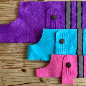 May include: Three felt dog vests in different sizes. The largest vest is purple, the middle vest is blue, and the smallest vest is pink. Each vest has a button and two reflective stripes.