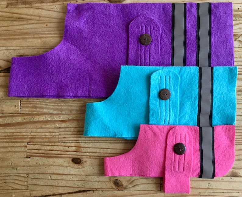 May include: Three felt dog vests in different sizes. The largest vest is purple, the middle vest is blue, and the smallest vest is pink. Each vest has a button and two reflective stripes.