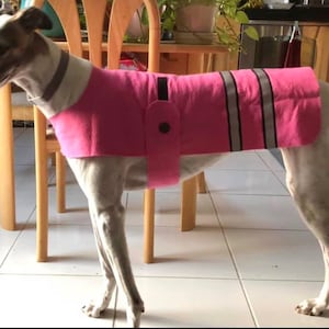 May include: A white dog wearing a pink fleece dog coat with reflective strips. The coat has a black button and a black strap.