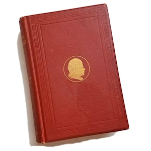 May include: A vintage, red hardcover book with gold accents. The cover features a gold profile portrait in a circular frame and a decorative border. The book's spine displays the title and author's name in gold lettering.