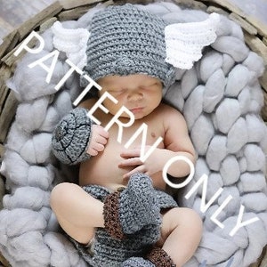 Könnte beinhalten: Eine graue gehäkelte Baby-Mütze mit weißen Flügeln, passende graue gehäkelte Booties und ein grauer gehäkelter Armwärmer. Das Baby trägt die Mütze und die Booties und liegt in einem Korb.