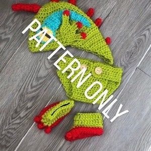 以下が含まれることがあります： 緑、赤、青のクロシェ編みベビー服。帽子、ズボン、ブーティが含まれています。帽子は角とスパイクのある恐竜のデザインです。ズボンは、前面にボタンが付いたシンプルなデザインです。ブーティは、赤いカフが付いたシンプルなデザインです。