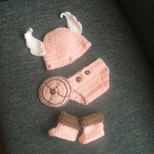 May include: A handmade, light pink crochet baby outfit set. The set includes a hat with white wings, a diaper cover with two brown buttons, and a pair of booties with brown cuffs. The set is designed for a newborn.