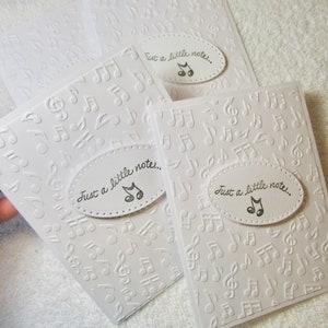 May include: A stack of white notecards with embossed musical notes. Each card has an oval label with the text "Just a little note..." and a musical note symbol. The cards are on a white background.