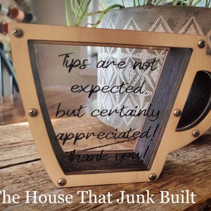 May include: A wooden cup-shaped tip jar with a clear acrylic front panel. The jar has a message that reads "Tips are not expected, but certainly appreciated! Thank you!" The House That Junk Built is written below the jar.
