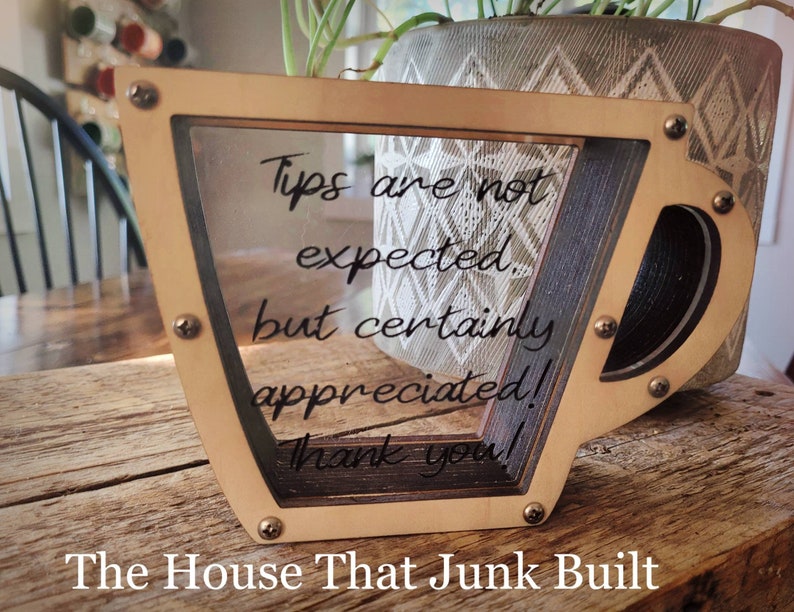 May include: A wooden cup-shaped tip jar with a clear acrylic front panel. The jar has a message that reads "Tips are not expected, but certainly appreciated! Thank you!" The House That Junk Built is written below the jar.