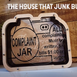 Può includere: Una scatola di legno a forma di macchina fotografica con un fronte di vetro. La scatola è etichettata "Complaint Jar" e ha il testo "Tutti i reclami devono essere scritti su banconote da $ 1,00 o più".