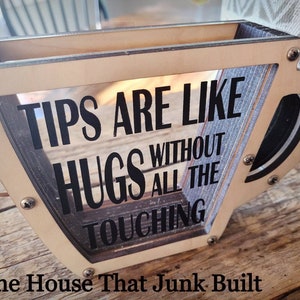 May include: A wooden cup-shaped tip jar with a clear front panel. The jar has a black sign that reads "Tips are like hugs without all the touching". The jar is sitting on a wooden surface. The House That Junk Built is written below the jar.