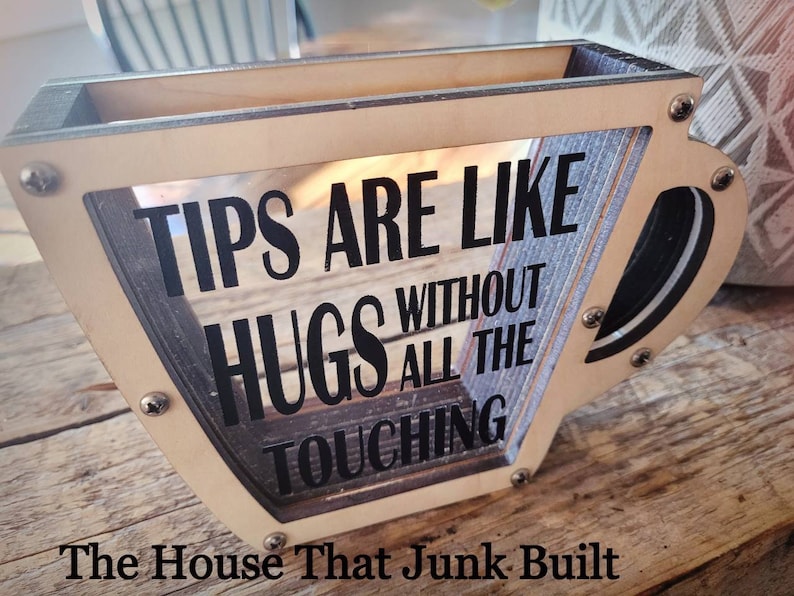 May include: A wooden cup-shaped tip jar with a clear front panel. The jar has a black sign that reads "Tips are like hugs without all the touching". The jar is sitting on a wooden surface. The House That Junk Built is written below the jar.