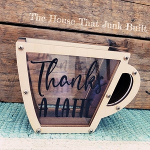 May include: A wooden coffee cup shaped money box with a clear plastic window. The cup has the words "Thanks a Latte" written on it in black.