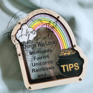 Könnte beinhalten: Holz-Sparschwein mit klarer Vorderseite, mit Regenbogen, Wolke und Goldtopf-Design. Der Text lautet "Things We Love: Mermaids, Fairies, Unicorns, Rainbows, and..." Das Wort "TIPS" wird auf dem Goldtopf angezeigt.