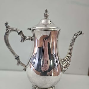 May include: A polished silver coffee pot featuring an ornate, curved handle, a decorative spout with intricate detailing, and a matching lid with a finial top. The body of the pot is rounded and supported by three detailed, scrolled feet.