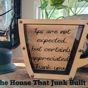 May include: A wooden cup-shaped tip jar with a clear plastic insert. The jar has a sign that reads "Tips are not expected, but certainly appreciated! Thank you." The jar is sitting on a wooden surface. The House That Junk Built is written below the jar.