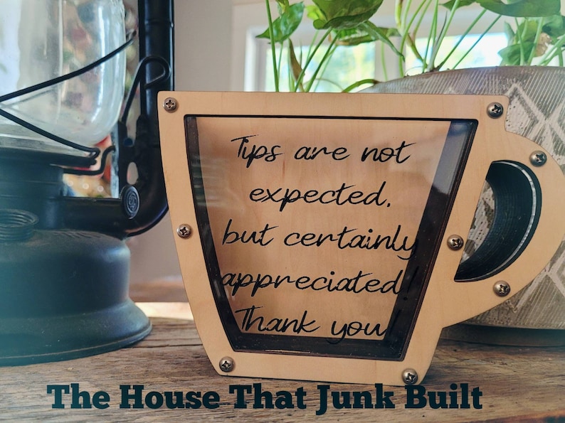 May include: A wooden cup-shaped tip jar with a clear plastic insert. The jar has a sign that reads "Tips are not expected, but certainly appreciated! Thank you." The jar is sitting on a wooden surface. The House That Junk Built is written below the jar.