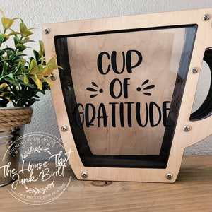 May include: A wooden cup-shaped money box with a glass front. The box is painted light brown and has a black outline. The words "CUP OF GRATITUDE" are printed in black on the front of the box.