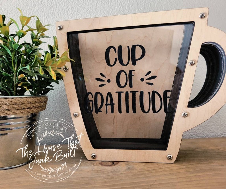 May include: A wooden cup-shaped money box with a glass front. The box is painted light brown and has a black outline. The words "CUP OF GRATITUDE" are printed in black on the front of the box.