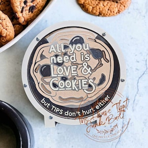 May include: A round wooden sign with a brown background and white text that reads "All you need is LOVE & COOKIES but TIPS don't hurt either". The sign is decorated with a cookie design and has a brown wooden frame.
