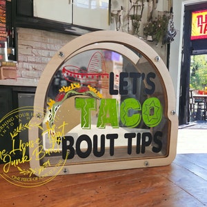 Peut inclure: Une enseigne en bois avec une fenêtre en plastique transparent, affichant le texte "Let's Taco Bout Tips". L'enseigne est décorée d'un graphique de taco vert et jaune et d'un sombrero rouge.
