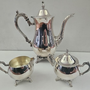May include: A three-piece silver-plated tea set consisting of a tall coffee pot with a lid, a sugar bowl with a lid, and an open creamer. Each piece features ornate, scrolled handles and feet with a polished, reflective metallic finish.