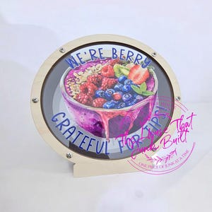 May include: A round wooden tip jar with a clear front, featuring a colorful illustration of a berry bowl. The text "WE'RE BERRY GRATEFUL FOR TIPS!" is displayed in blue. The jar is designed with a light wood finish.