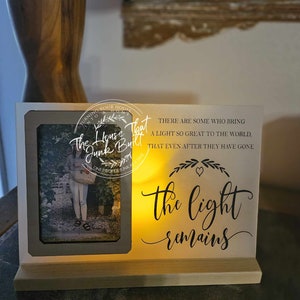 Puede incluir: Un marco de fotos de madera blanco con una vela encendida en su interior. El marco tiene una inscripción en negro que dice "Hay quienes traen una luz tan grande al mundo, que incluso después de que se hayan ido, la luz permanece".