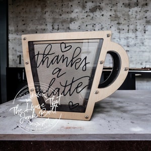 May include: A wooden coffee cup shaped box with a glass front. The box has a black painted handle and the words "thanks a latte" written on the glass. The box is sitting on a light grey surface.