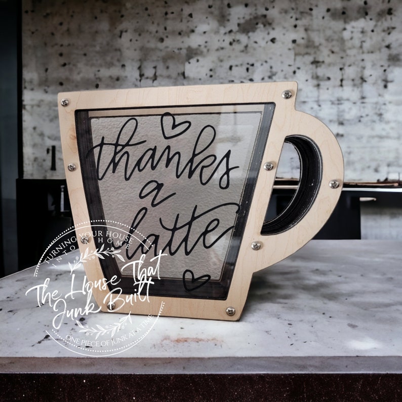 May include: A wooden coffee cup shaped box with a glass front. The box has a black painted handle and the words "thanks a latte" written on the glass. The box is sitting on a light grey surface.