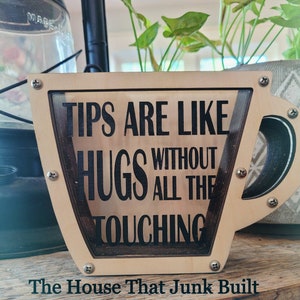 May include: A wooden sign shaped like a coffee mug with the text "Tips are like hugs without all the touching". The sign is painted in a light brown colour and has a dark brown border. The text is in black.